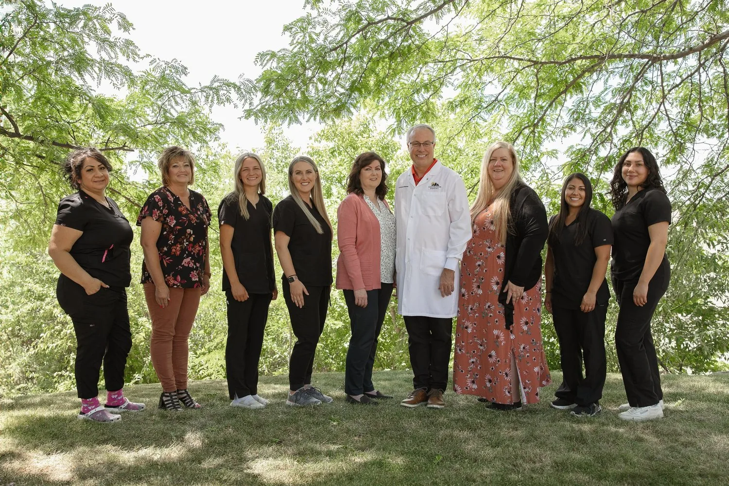 Dental team including a dentist in a white lab coat and staff in scrubs standing outdoors under trees.